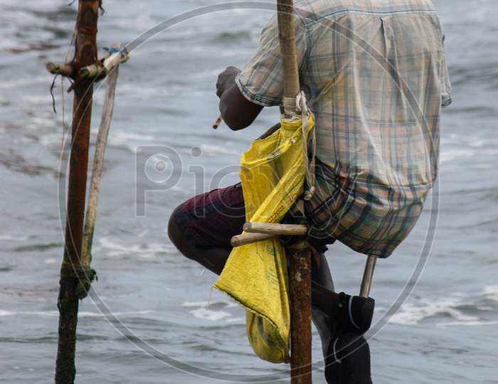 Stilt Fisherman With His Wooden Rod Facing Back To The Camera With His Yellow Pocket On The Pole, Fishing In A Traditional Unique Method In Sri Lankan Culture, Sunny Bright Evening On The Beach.