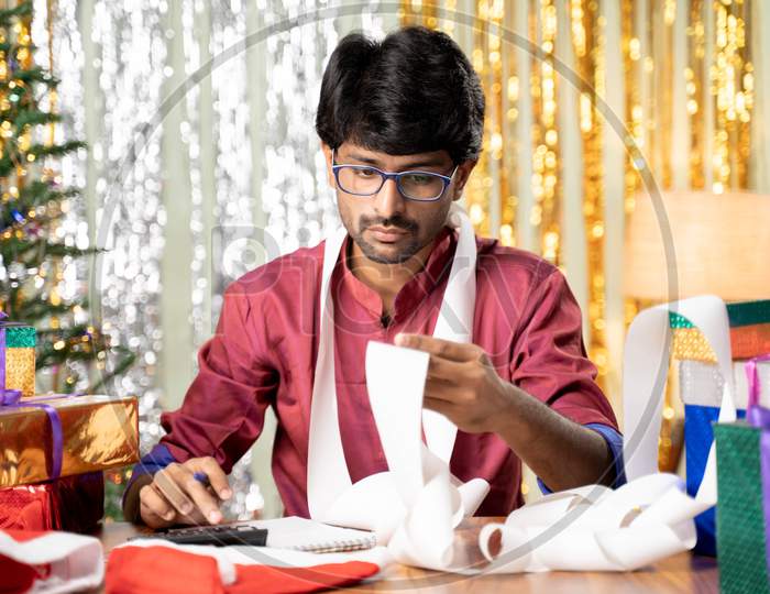 Young Man Busy In Calculating Holyday Expenses After Christmas Or New Year 2021 Holiday Celebration Showing With Decorated Background With Gift In Front