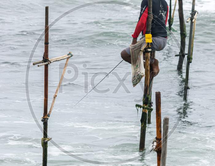 Stilt Fisherman With His Wooden Rod Facing Side With His Pocket On The Pole To Fill With Fish, Fishing In A Traditional Unique Method In Sri Lankan Culture, Wearing A Cap To Prevent Sunburns.