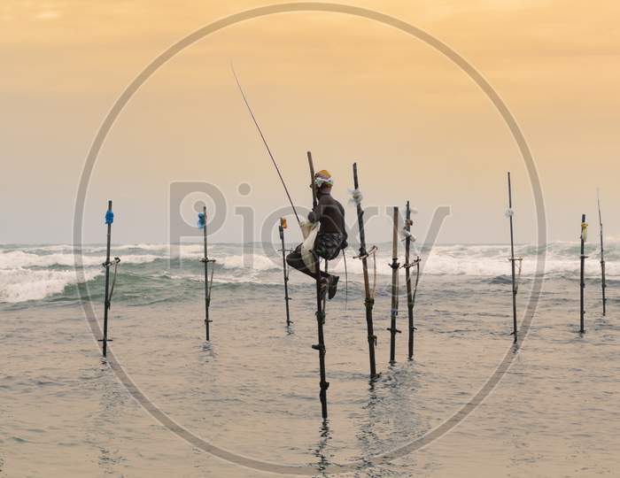 Stilt Fisherman Sitting In His Pole With A Wooden Fishing Rod In His Hands In The Sunset Evening. Ocean Waves Crash Behind Him In The Background.