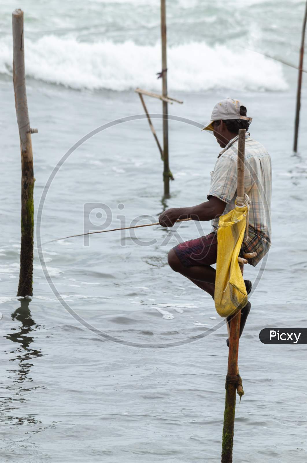 Stilt Fisherman With His Wooden Rod Facing Side To The Camera With His Yellow Pocket On The Pole, Fishing In A Traditional Unique Method In Sri Lankan Culture, Sunny Bright Evening On The Beach.