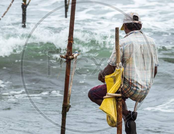 Stilt Fisherman With His Wooden Rod Facing Back To The Camera With His Yellow Pocket On The Pole, Fishing In A Traditional Unique Method In Sri Lankan Culture, Sunny Bright Evening On The Beach.