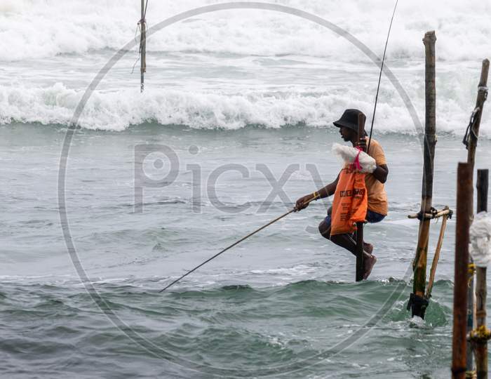 Weligama, Southern Province / Sri Lanka - 07 26 2020: Stilt Fishermen With His Wooden Rod Facing Side To The Camera With His Orange Pocket, Fishing In A Traditional Unique Method In Sri Lankan Culture