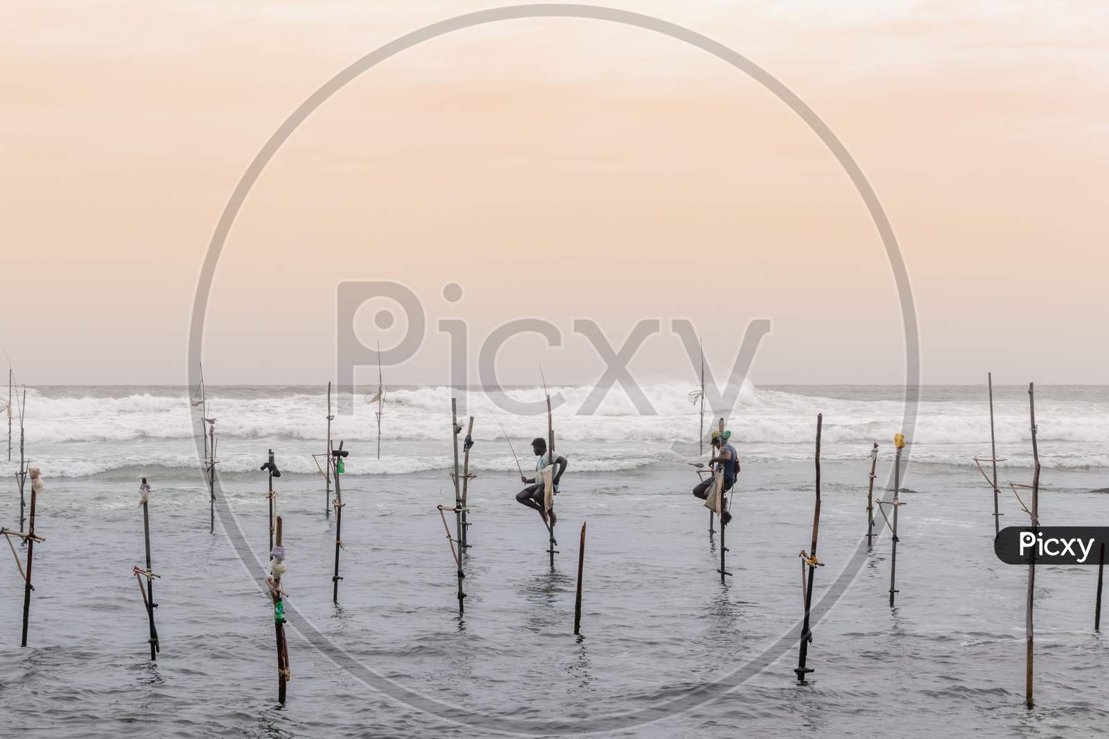 A Couple Of Stilt Fishermen Sitting On Their Poles With Wooden Fishing Rods In Their Hands In The Sunset Evening. Ocean Waves Crash Behind Them In The Background With An Orange Yellowish Sky.