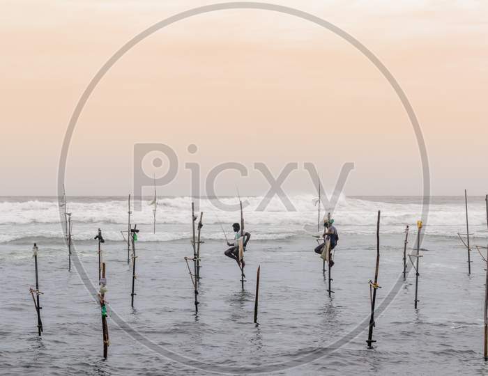 A Couple Of Stilt Fishermen Sitting On Their Poles With Wooden Fishing Rods In Their Hands In The Sunset Evening. Ocean Waves Crash Behind Them In The Background With An Orange Yellowish Sky.