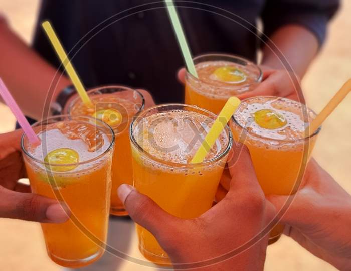 Five friends together in a circle holding their orange cocktail with ice and straw. They are celebrating their friendship or success in the beach. They are having cheers