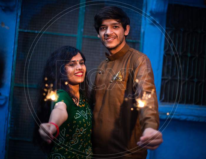 Happy Cheerful Young Indian Couple Celebrating Diwali Festival Enjoying Sparklers Firecrackers As Part Of The Celebration And Holidays, Wearing Traditional Outfits