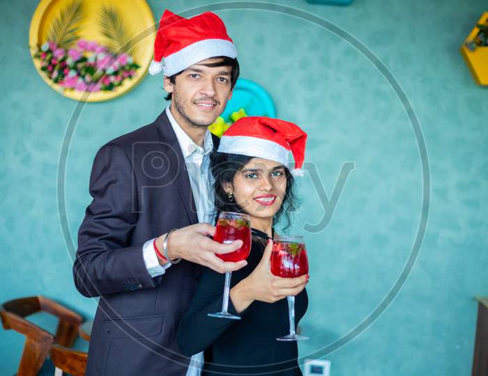 Happy Cheerful Young Couple Wearing Santa Hats Holding Red Cocktail Drink Glasses Celebrating Christmas Looking At Camera, Copy Space.