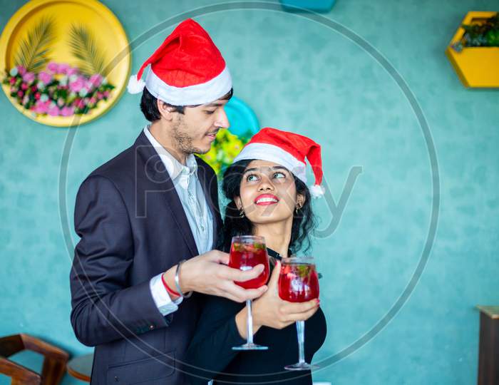 Happy Young Couple Wearing Santa Hats Celebrating Christmas Together While Holding Red Cocktail Drink Glasses Looking At Each Other At Home, Copy Space. Festivity Concept.