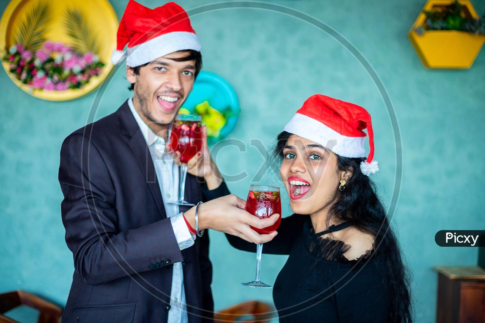 Happy Young Couple In Santa Hats Celebrating Christmas Together While Having Fun With Red Cocktail Drink Glasses Love, Bonding, Copy Space.