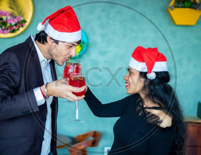 Happy Young Indian Couple In Santa Hats Celebrating Christmas Together While Having Fun With Red Cocktail Drink Glasses Love, Bonding,