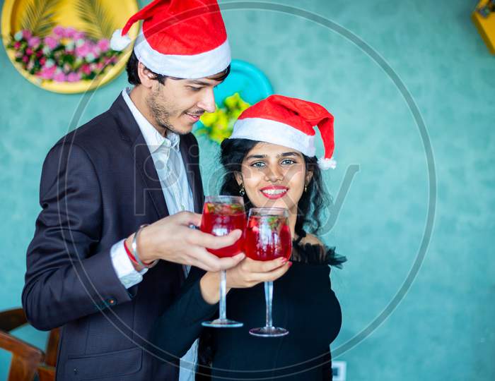 Christmas Celebration, Happy Young Couple Wearing Santa Hats Cheering Red Cocktail Drink Glasses At Home, Copy Space. Love, Bonding, Party Concept.