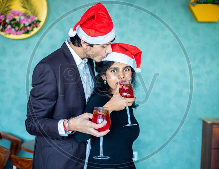 Young Couple In Santa Hats Celebrating Christmas Together Holding Red Cocktail Drink Glasses Boyfriend Kisses On Her Girlfriends Forehead, Love, Bonding Copy Space.
