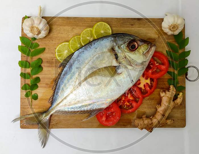 Close Up View Of Fresh Malabar Trevally Fish Decorated With Curry Leaves , Tomato,Lemon Slice And Herbs On A White Background.