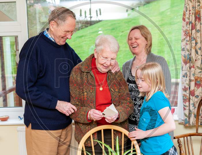 Three Generations Of A Family Laughing While An Elderly Lady Reads A Birthday Card