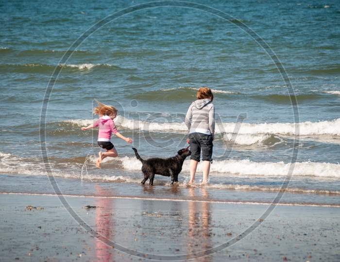 A Mother And Daughter With A Black Dog Jump Over The Breaking Waves On A Beach