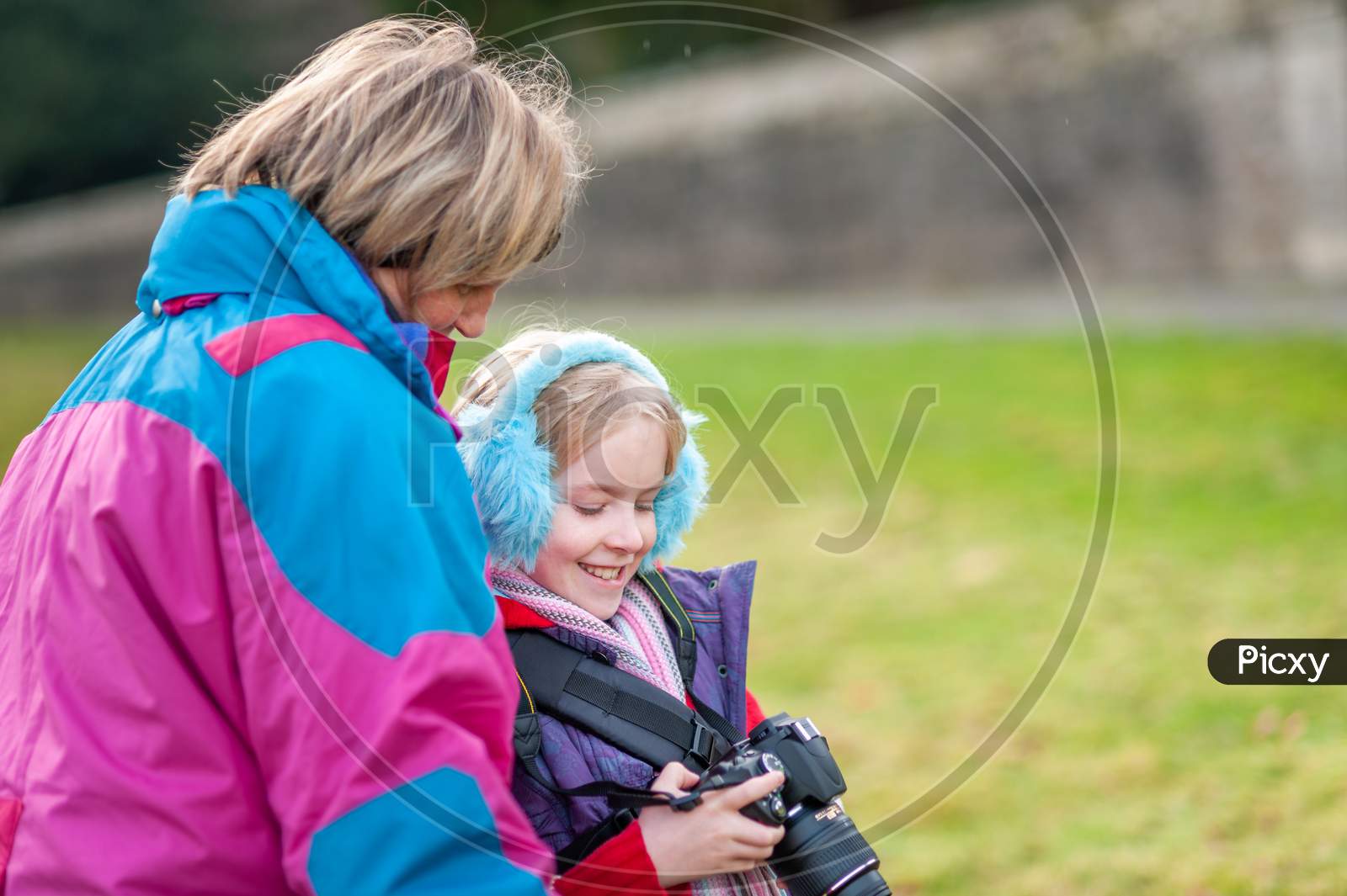 Happy Young Girl Holding A Camera And Wearing Blue Ear Muffs Shows Camera To Mother