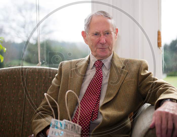 A Well Dressed Elderly Man Sat In Front Of A Bright Window