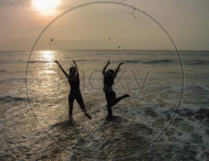 Silhouette Of Two Happy Women On African Beach Throwing Sand In The Air At Sunset.