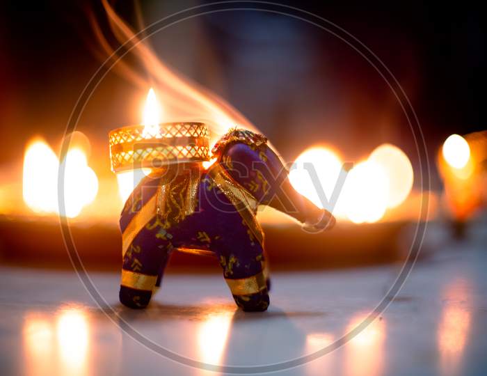 Small Elephant Incense Holder With Smoke Coming Out And An Out Fo Focus Background With Diya Oil Lamps Lit Behind It With Flickering Flames