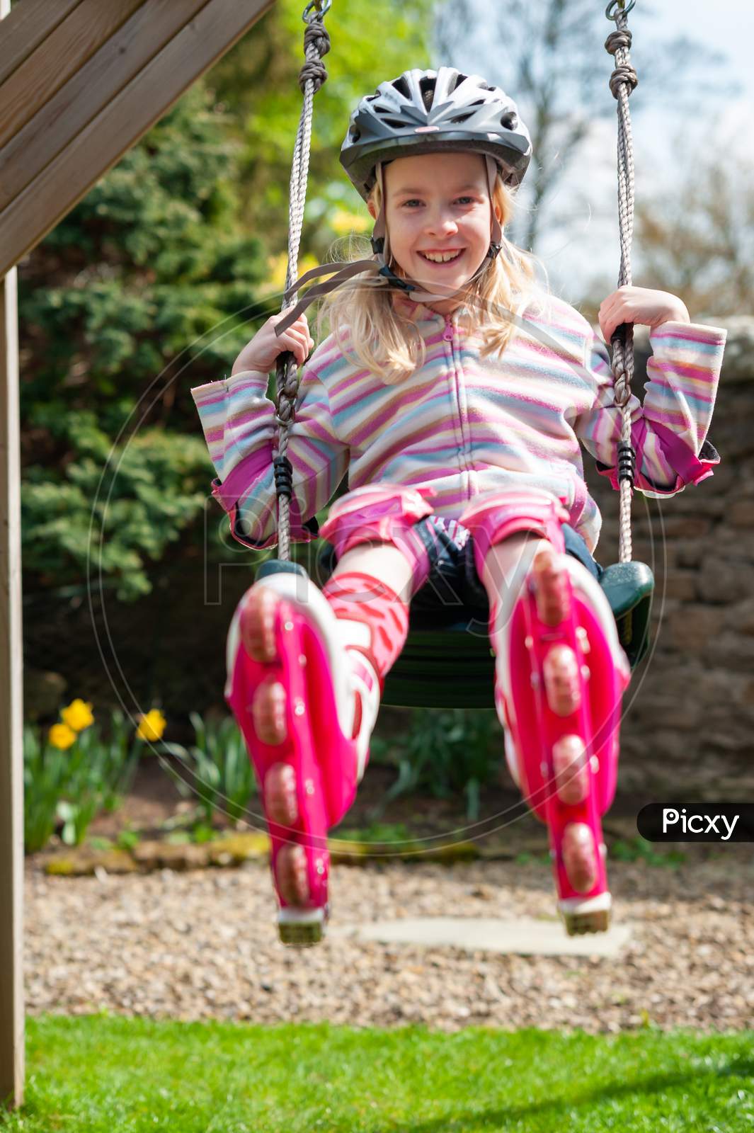 A Smiling Young Blonde Girl Wearing Roller Blades And Helmet While Swinging On Garden Swing