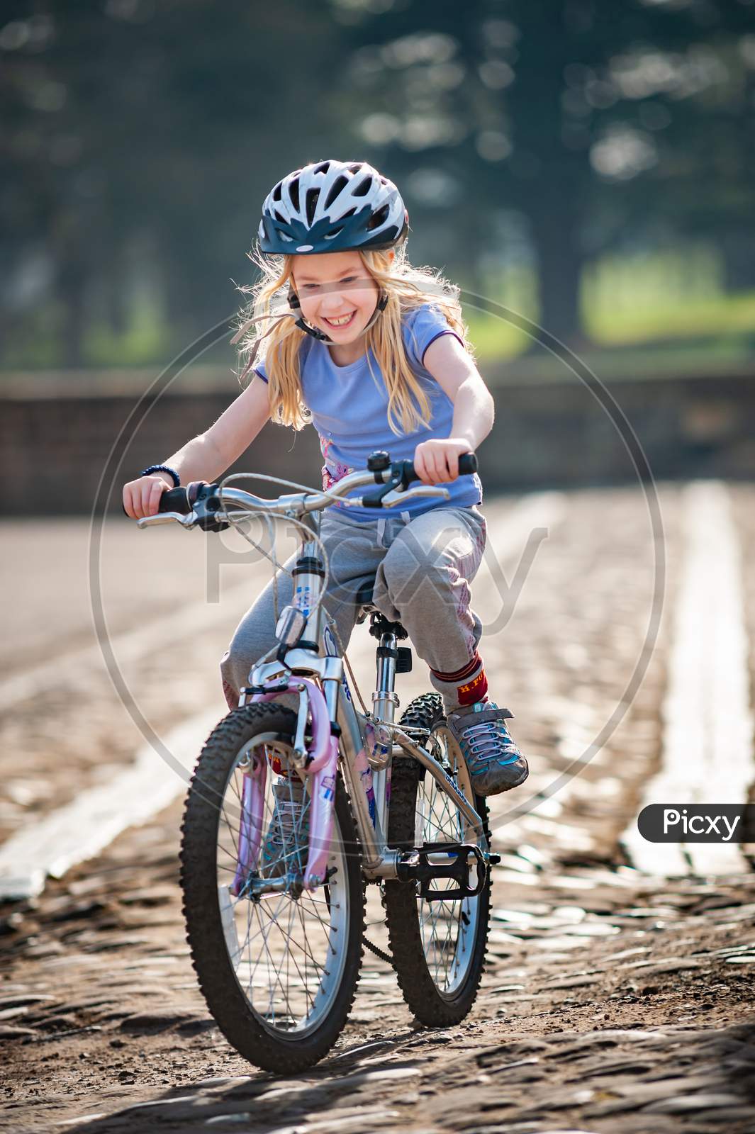 A Very Happy Young Blonde Girl Rides A Bike Along A Cobbled Path Towards Camera In Golden Sunlight