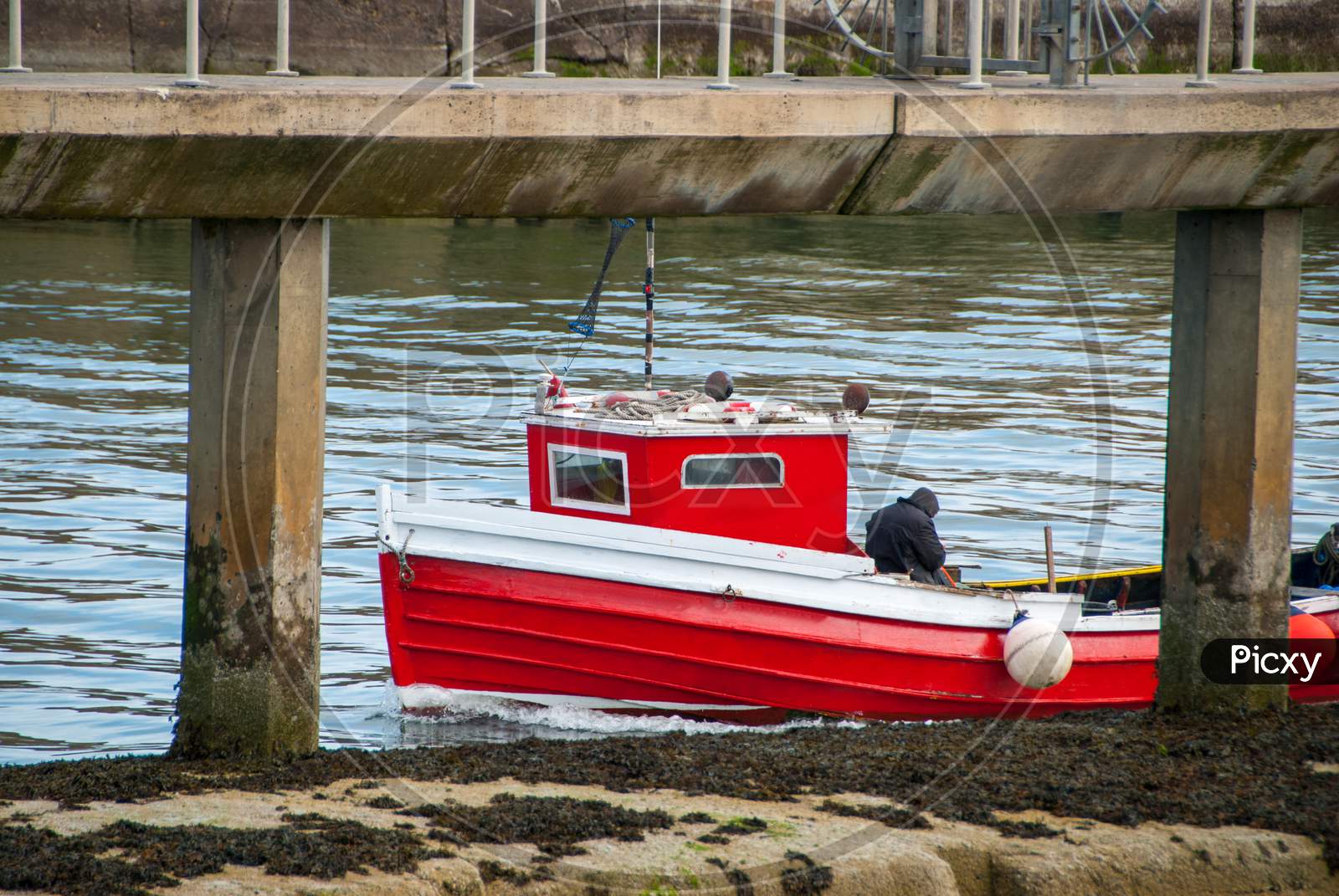 A Small Red Fishing Boat Between The Posts Of A Pier On A Calm Day