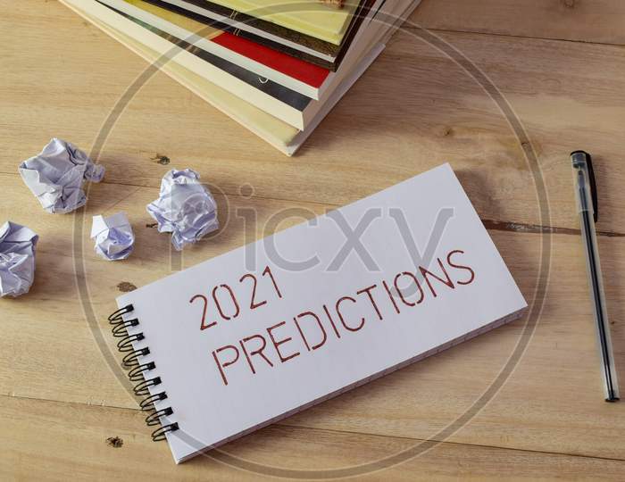 2021 PREDICTIONS handwritten in notepad on wooden desk with pen, books and crumbled paper balls.