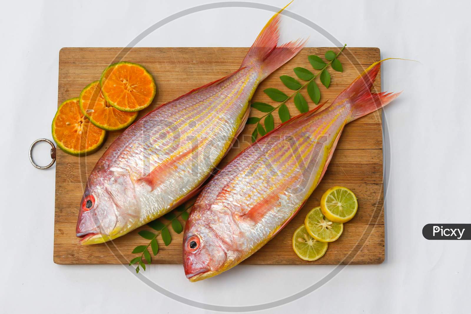 Top View Of View Of Fresh Pink Perch (Thread Finned Bream) Decorated With Lemon Slice,Orange Slice And Curry Leaves On A Wooden Pad,White Background,Selective Focus.
