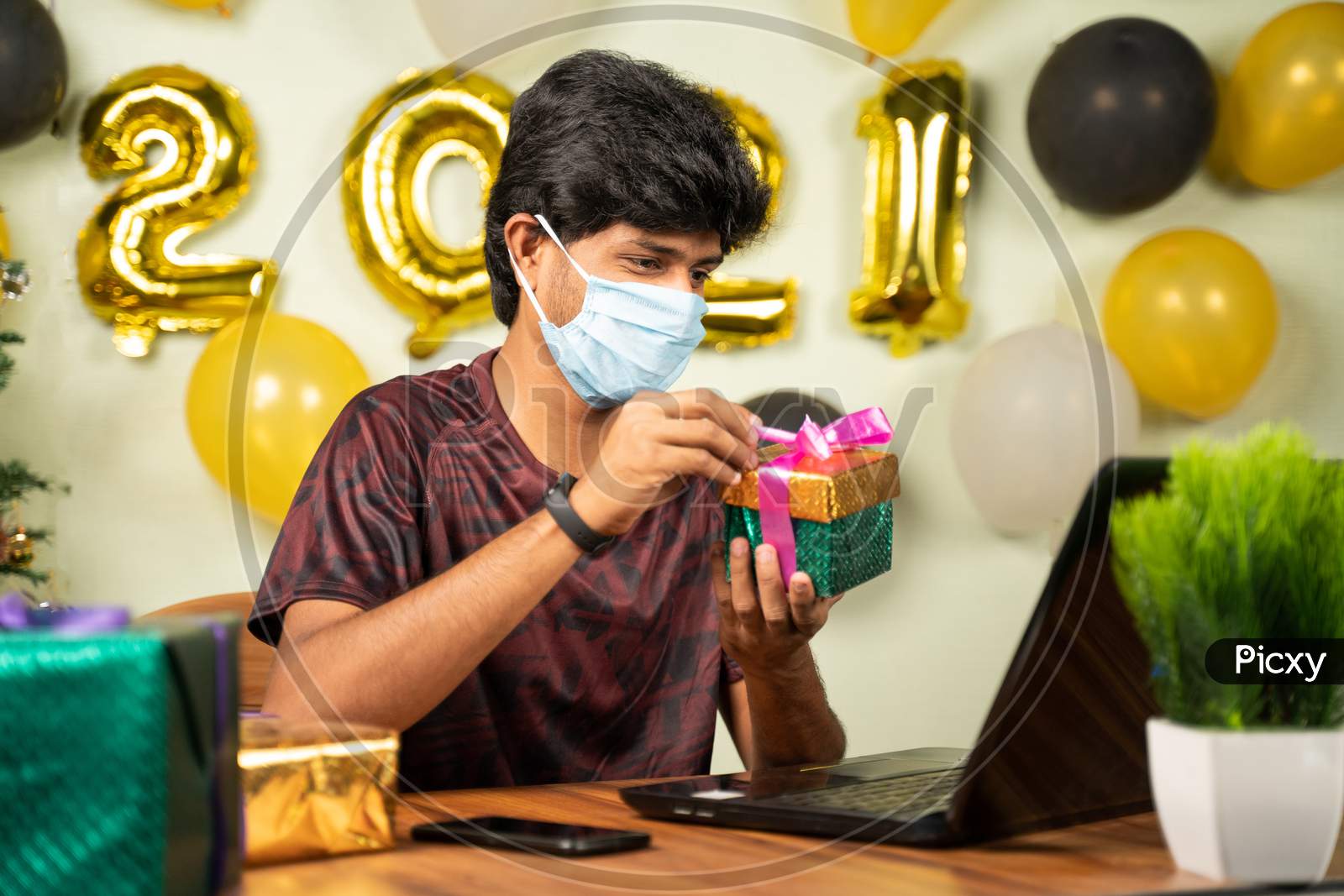 Young Man With Medical Mask Opening Gift Box Over Video Call On Laptop With 2021 New Year Decorated Background - Concept Of Distant Xmas Or Holiday Celebration Due To Covid-19 Or Coronavirus Pandemic.