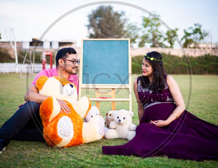 Young Pregnant Asian Indian Couple Looking At Each Other Sitting In A Garden With Lots Teddy Bear Toys And Blank Chalkboard To Write Message. Pregnancy Concept.