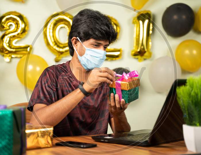 Young Man With Medical Mask Opening Gift Box Over Video Call On Laptop With 2021 New Year Decorated Background - Concept Of Distant Xmas Or Holiday Celebration Due To Covid-19 Or Coronavirus Pandemic.