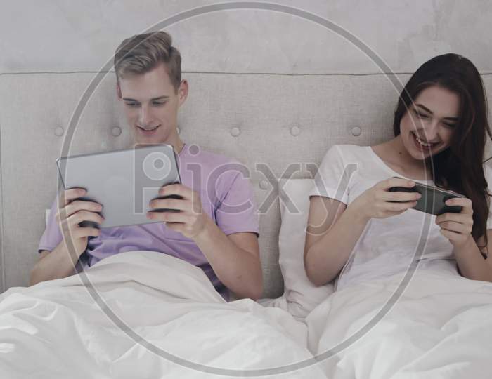 Couple Playing Mobile Games In Bed. Couple Playing On Tablet And Phone. Couple In Bed Using Phone And Tablet For Games. Morning Time. Shot On Red