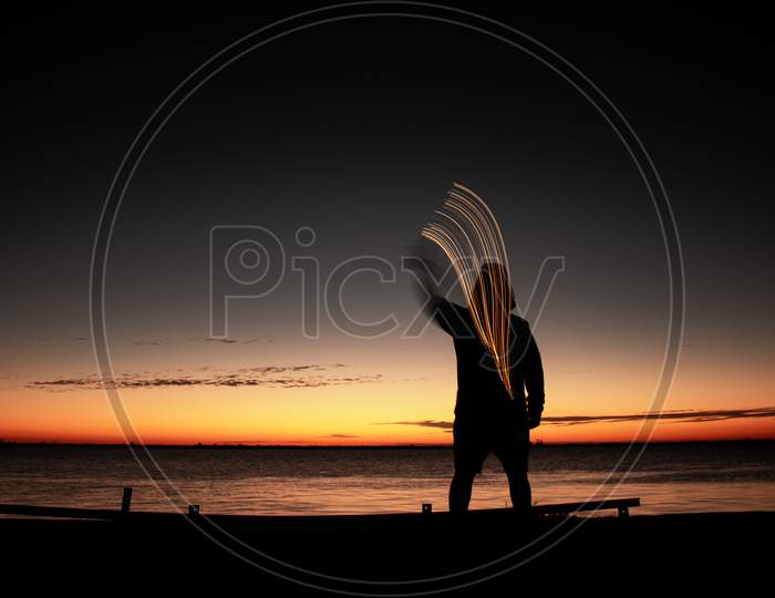 Human Figure Moving A Light During Sunset. Light Painting On The Edge Of The River.