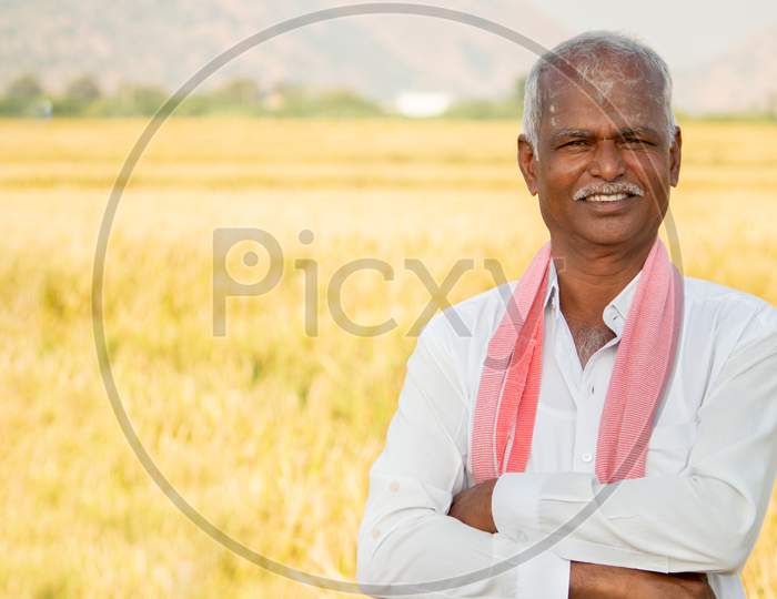 Portrait Of Confident Smiling Indian Farmer With Arms Crossed Standing In Front Of Agriculture Farmland Looking Camera With Copy Space.
