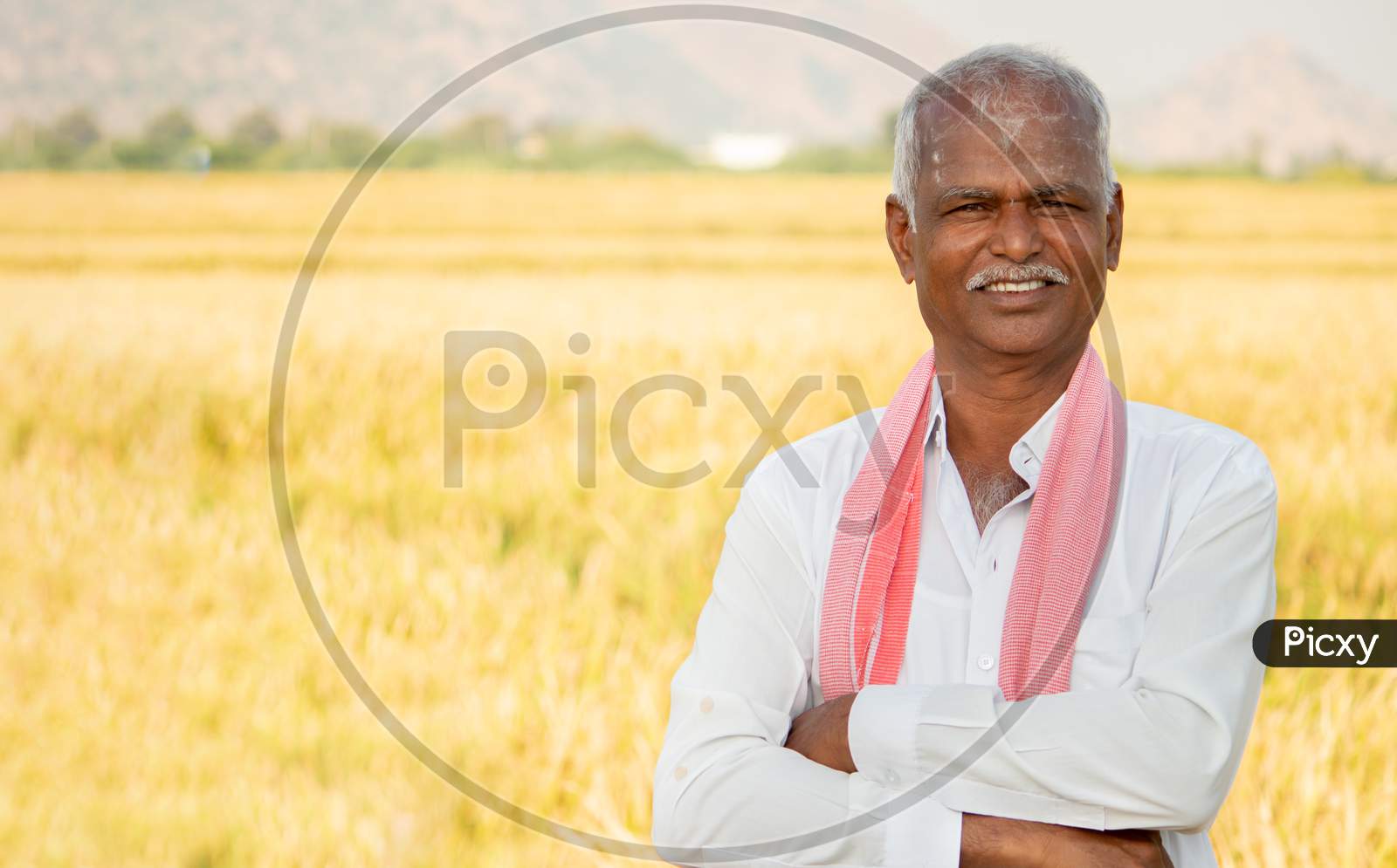 Portrait Of Confident Smiling Indian Farmer With Arms Crossed Standing In Front Of Agriculture Farmland Looking Camera With Copy Space.