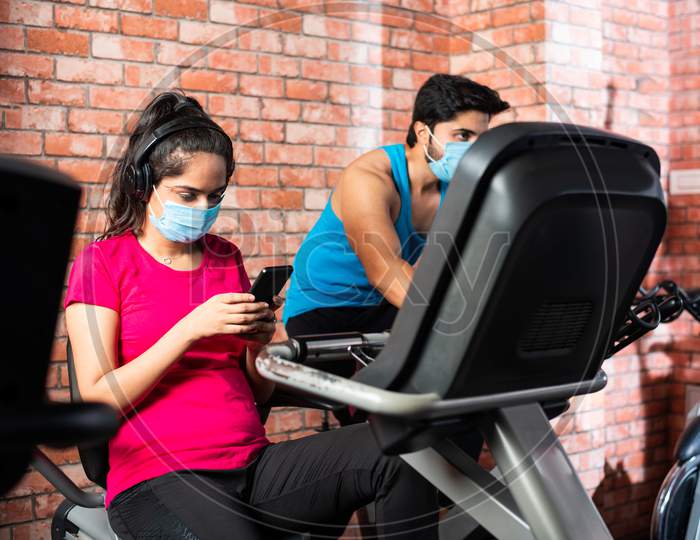 Indian Young Couple Wears Face Mask While Riding On Fitness Bike In The Gym