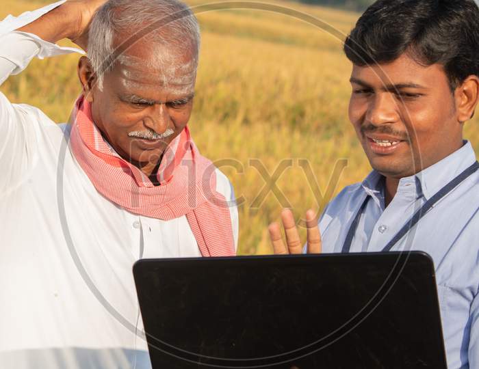 Confused Indian Farmer Scratching His Head While Banker Or Corporate Government Officer Discussing On Laptop About Crop Yield, Credit And Loan Subsidy At Agriculture Farmland During Hot Sunny Day