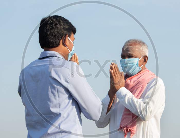 Low Angle View, Farmer Greeting To Banker Or Corporate Government Officer By Doing Namaste While Both Worn Face Mask Due To Coronavirus Covid-19 Pandemic Meeting Near Agriculture Farmland.