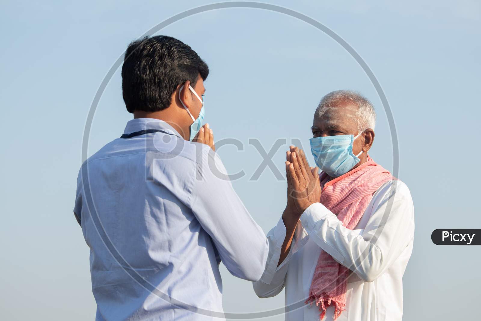 Low Angle View, Farmer Greeting To Banker Or Corporate Government Officer By Doing Namaste While Both Worn Face Mask Due To Coronavirus Covid-19 Pandemic Meeting Near Agriculture Farmland.