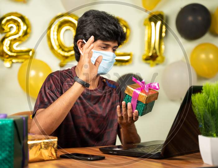 Selective Focus On Gift Box, Young Man With Medical Mask Got Gift Box Over Video Call On Laptop With 2021 New Year Background - Concept Of Distant Holiday Celebration Due To Covid-19 Or Coronavirus.