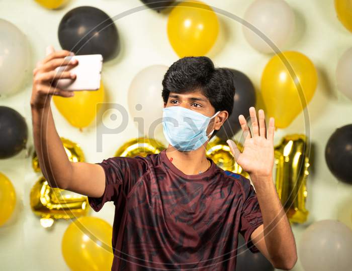 Young Man With Medical Mask Busy Making Video Call On Mobile Over 2021 New Year Decorated Background - Concept Of Quarantine Holyday Celebration Due To Pandemic