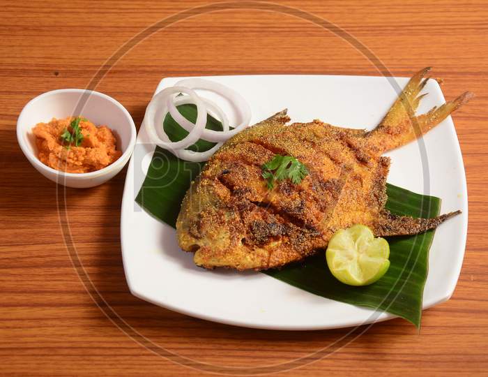 Pomfret Fish Tawa Fry Garnished With Lemon And Onion In A White Ceramic Plate With Wooden Background,Selective Focus