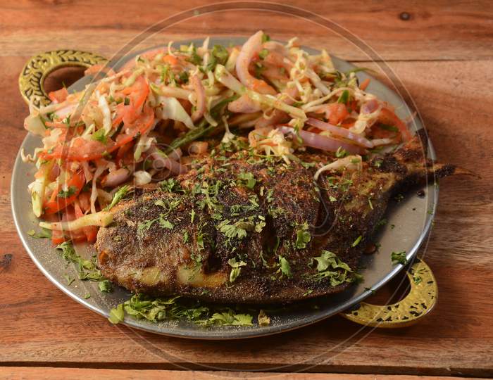 Pomfret Fish Tawa Fry Which Is Well Garnished With Tomato,Cabbage, Lemon And Onion Served In Wooden Background,Selective Focus