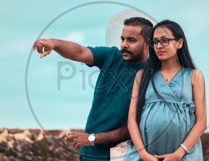A Young Couple, Husband Pointing At Something In The Distance, A Beautiful Pregnant Lady With A Cute Smile, Standing From Belly High Portrait In Natural Light, And Great Weather Blue Skies.