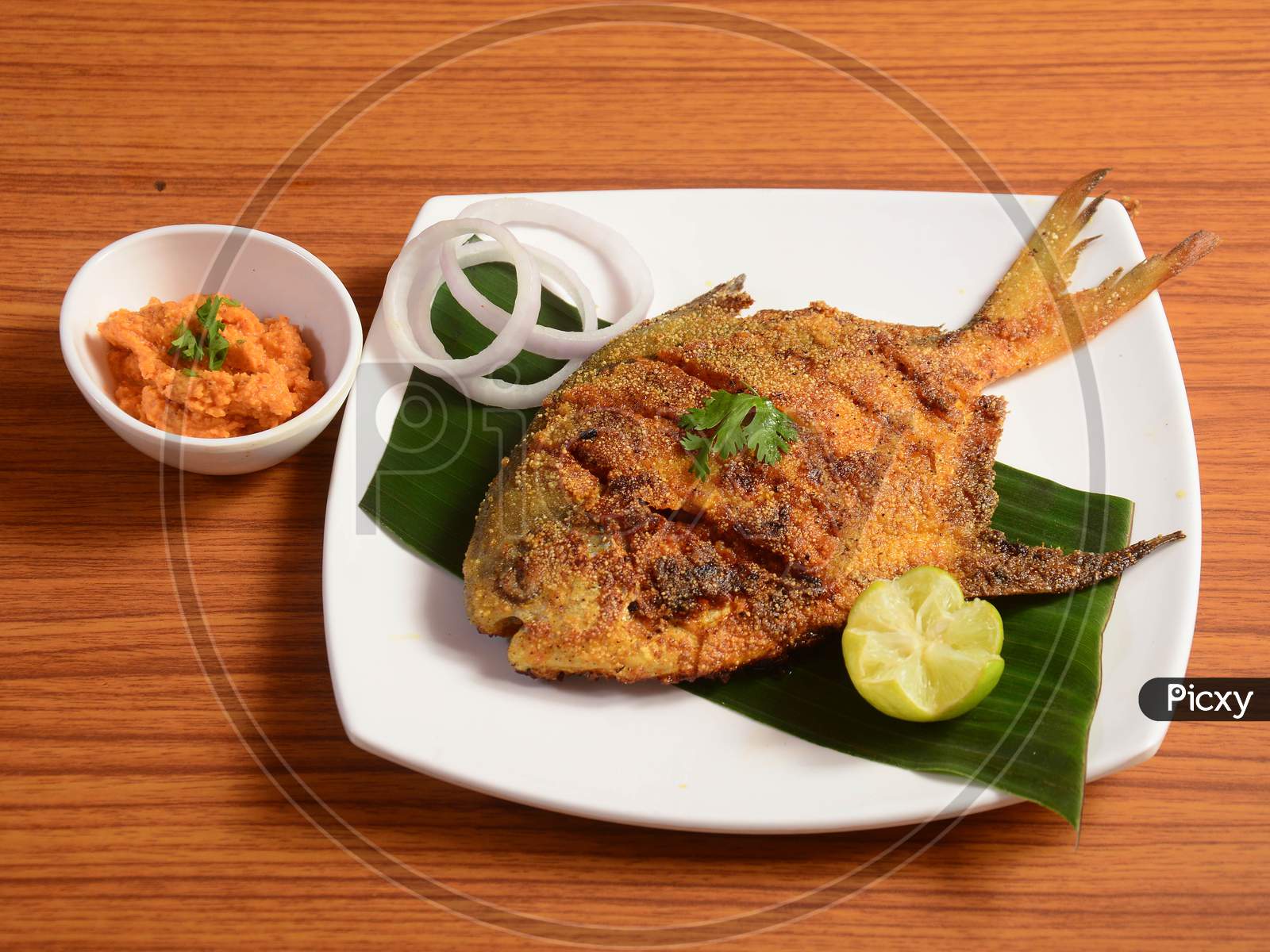Pomfret Fish Tawa Fry Garnished With Lemon And Onion In A White Ceramic Plate With Wooden Background,Selective Focus