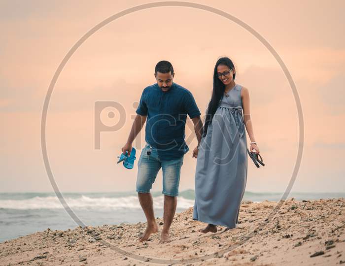 A Romantic Young Couple Expecting A Baby, Walk On The Beach Holding Hands Together In The Evening Sunset With Slippers In Hand.