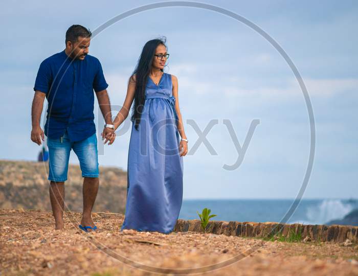 Holding Hands And Casually Walking Forward As Looking At The Sea In The Galle Fort Pathway, A Young Pregnant Couple Relaxing In The Evening Time, Blue Sky, And Ocean In The Background.