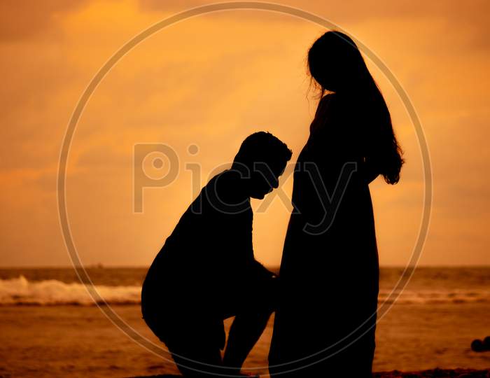Husband On His Knees In The Beach Evening Sunset Silhouette, Kissing Pregnant Wife'S Belly, Parenthood Concept.