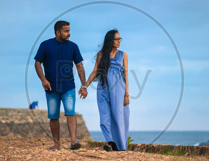 Holding Hands And Casually Walking Forward As Looking At The Sea In The Galle Fort Pathway, A Young Pregnant Couple Relaxing In The Evening Time, Sunlight Hitting Their Faces.